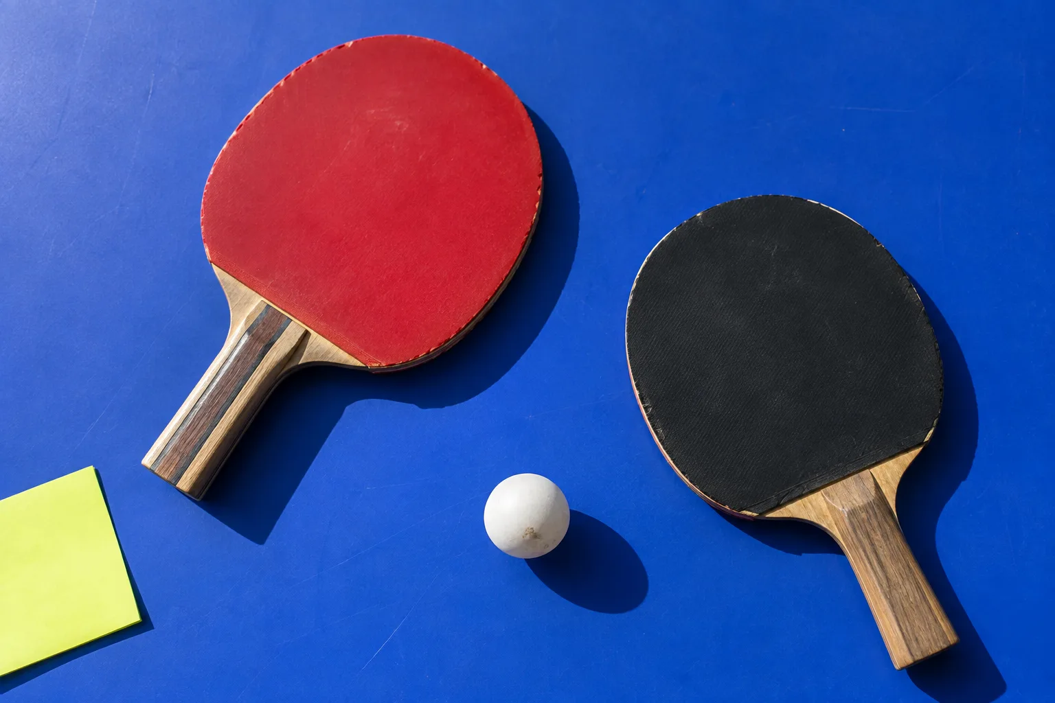 Two worn table-tennis paddles and a ball on a blue table, beside a blank lime note.